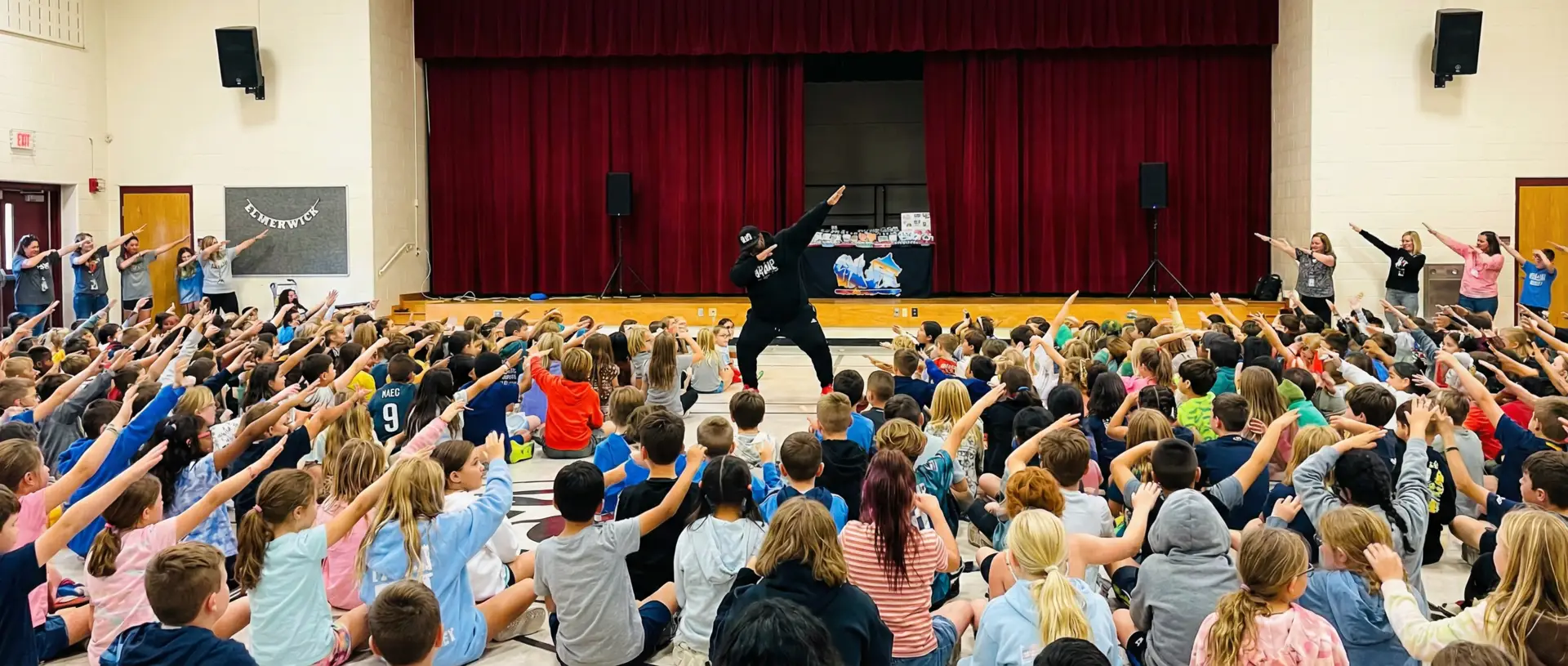 Large group of elementary students sitting on gymnasium floor with hands raised during the Respect Spells Hip Hop character education assembly.